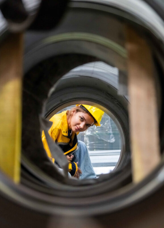 Portrait of Female Engineer examining quality of metal steel pipes by looking inside the tube at factory warehouse.