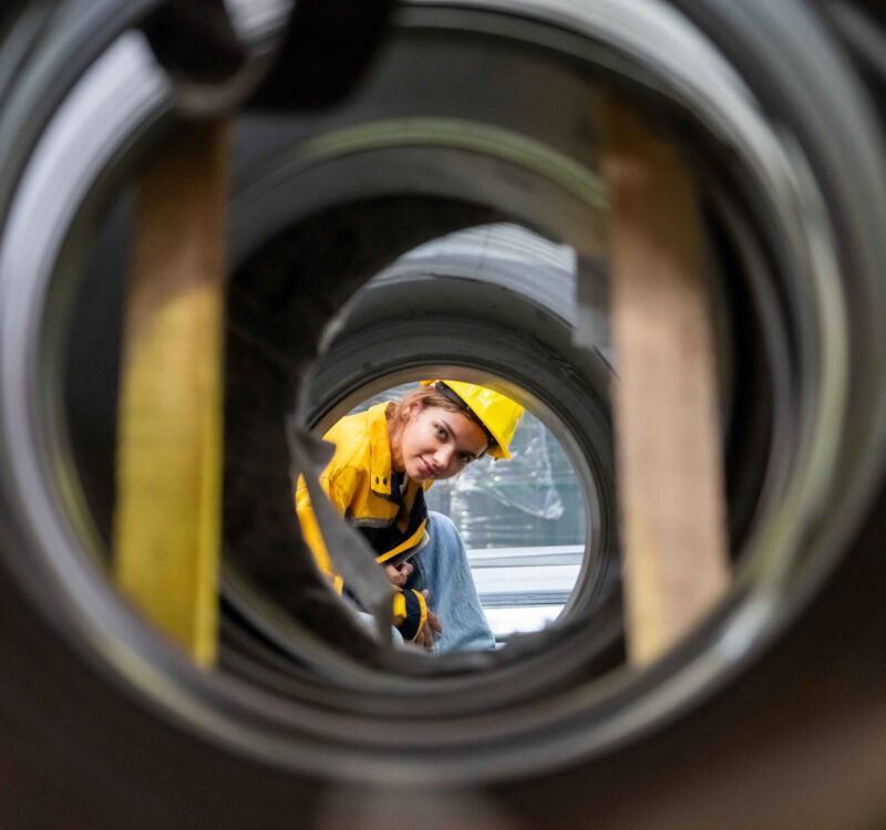 Portrait of Female Engineer examining quality of metal steel pipes by looking inside the tube at factory warehouse.